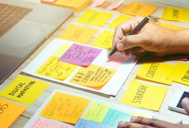 Image of person holding pen over multi-colored sticky notes on a desk