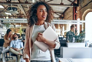 Female business professional holding laptop and smiling while sitting on desk and looking out the window