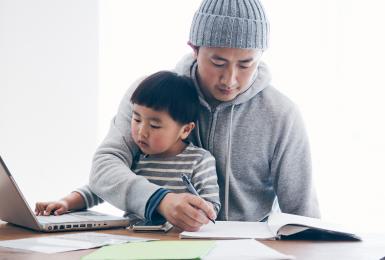 Japanese man in casual clothes writing a document and his son using a laptop on the desk. He's working and doing childcare at home.