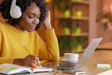 Smiling female student with wireless headset studying online, using laptop at cafe, taking notes, copy space