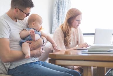Dad holds their daughter at the kitchen table while Mom works on her laptop next to him.