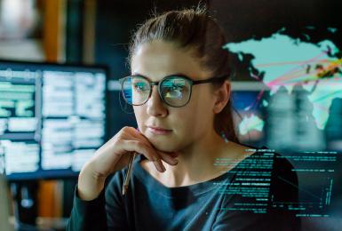 Stock image of a young woman, wearing glasses, surrounded by computer monitors in a dark office. In front of her there is a see-through displaying showing a map of the world with some data.