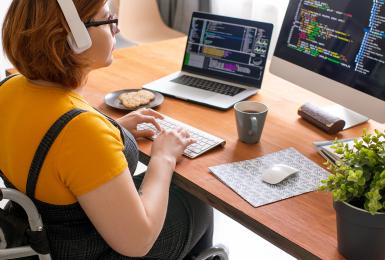 Female freelance IT tech in modern headphones sitting in wheelchair and using computers while coding