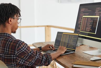 African-American IT developer typing on keyboard with black and orange programming code on computer screen and laptop in contemporary office interior