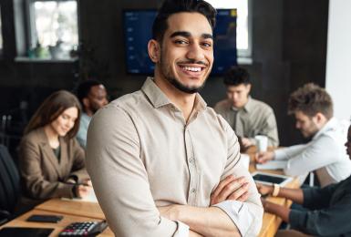 Successful businessman smiles at camera with colleagues in background meeting.