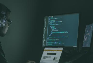 Man with headphones sits in a dark room in front of 3 computer screens