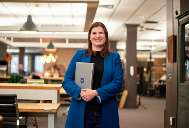 Woman standing in library holding laptop smiling portrait