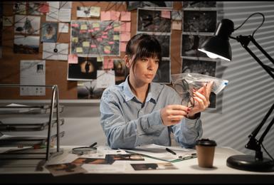  investigator examining evidence in a bag at a desk surrounded by case photos and a crime board