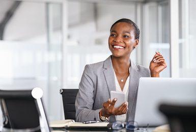smiling businesswoman in a gray blazer sits at her desk holding a smartphone, with a laptop open in front of her in a bright office setting.