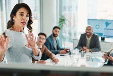A woman in a light blue sweater speaks and gestures expressively at the head of a conference table 