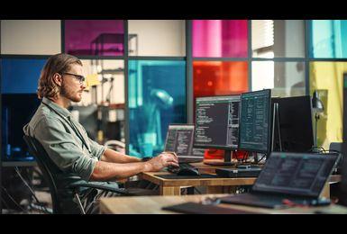 A software developer with glasses works at a multi-monitor desktop setup