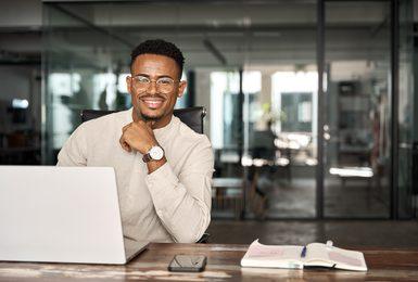A young professional man wearing glasses sits at a desk with a laptop