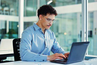 A young man with curly hair and glasses sits at a desk in a modern office, typing on a laptop.