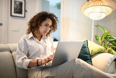A young woman with curly hair and glasses works on a laptop 