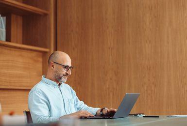 A middle-aged man with glasses and a short beard sits at a desk working on a laptop in a warmly lit office with wood-paneled walls.