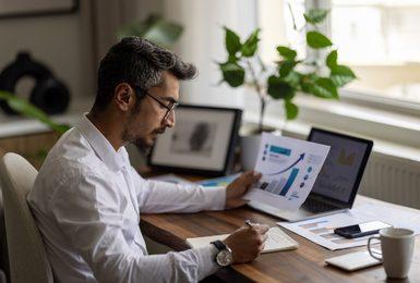 A man in a white dress shirt and glasses sits at a wooden desk reviewing financial charts 