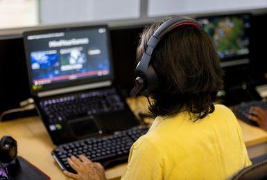 A person wearing a headset works at a laptop displaying video game design software, with additional monitors visible in the background.