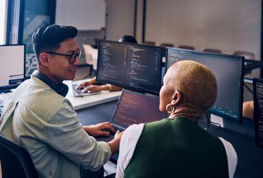 Two students review code together at a laptop in a computer lab, with monitors displaying programming windows in the background.