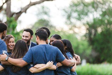 A diverse group of colleagues in matching t-shirts stand in a huddle outdoors with arms around each other, representing community and social connection.