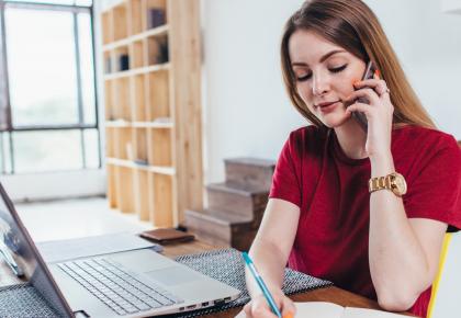 Female professional in front of laptop on kitchen table, talking on cellphone and taking notes