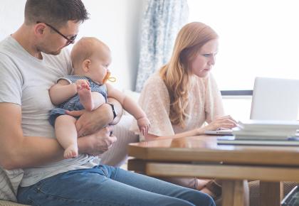 Dad holds their daughter at the kitchen table while Mom works on her laptop next to him.
