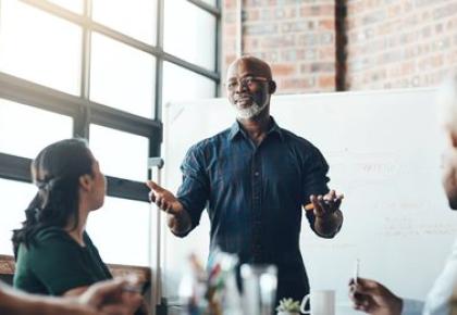 man with glasses and a gray beard stands at a whiteboard gesturing with both hands while leading a team discussion