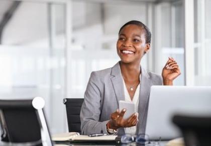 smiling businesswoman in a gray blazer sits at her desk holding a smartphone, with a laptop open in front of her in a bright office setting.