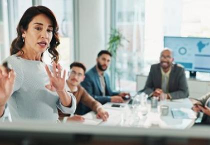 A woman in a light blue sweater speaks and gestures expressively at the head of a conference table 