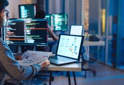 A software developer sits at a desk reviewing a printed UI wireframe
