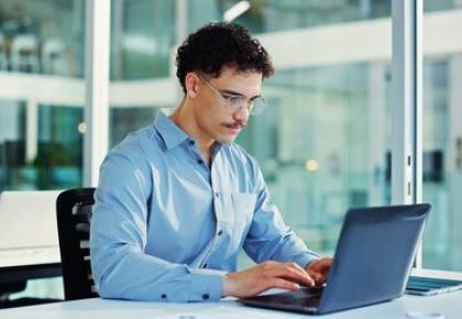 A young man with curly hair and glasses sits at a desk in a modern office, typing on a laptop.