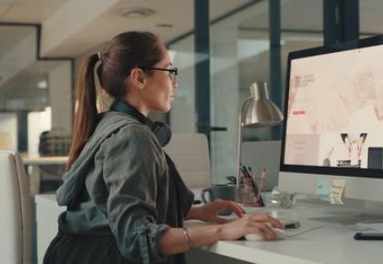 A woman with headphones around her neck sits at a desktop computer in a modern office, browsing a website on a large monitor.
