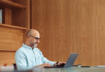 A middle-aged man with glasses and a short beard sits at a desk working on a laptop in a warmly lit office with wood-paneled walls.