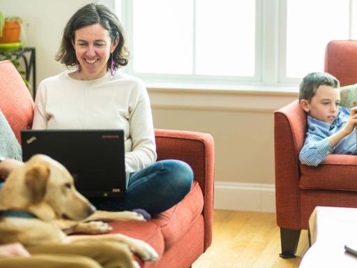 Woman studying on laptop on couch at home with husband and dog while son is reading nearby