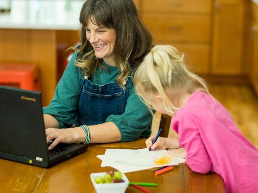 Mother on laptop happily doing accounting school work on laptop while young daughter colors next to her. 