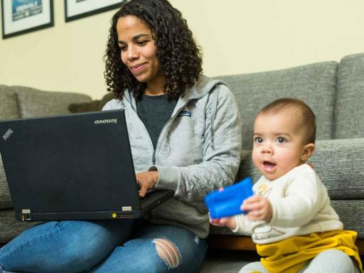 Female student working on laptop with her baby playing with a block nearby