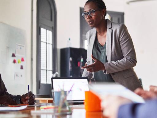 Woman of color standing, leading a meeting with a table full of employees