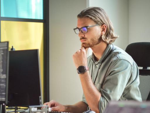 young man looking sitting at a desk looking intently at two monitors