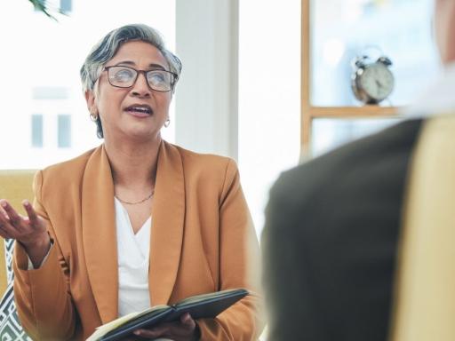 woman seated in chair facing seated man we see his back she is talking to him with a notepad in one hand