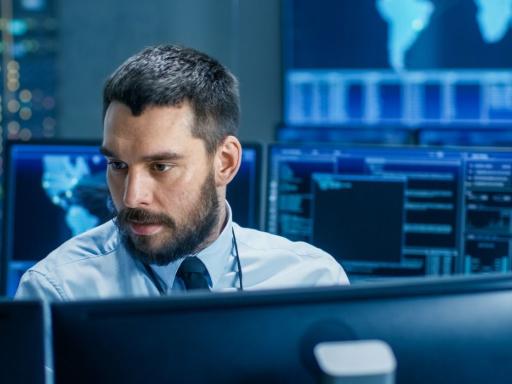 man facing monitors with a variety of monitors displaying different code behind him