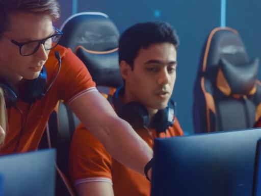 Three gamers, two female one male, behind their monitors with an esports coach pointing to the male's monitor