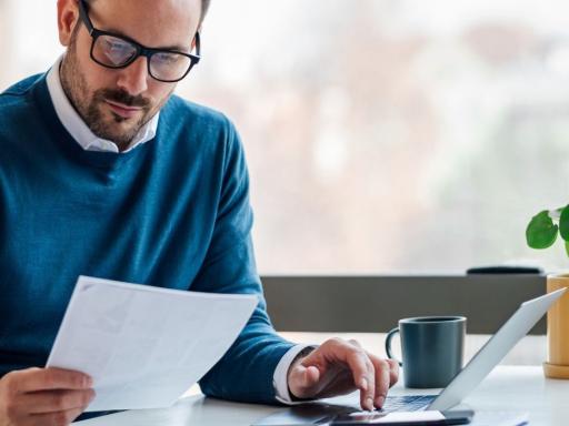 man with glasses and in blue sweater looking at a paper while typing on a laptop at his desk