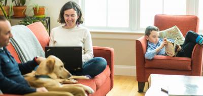 Woman studying on laptop on couch at home with husband and dog while son is reading nearby