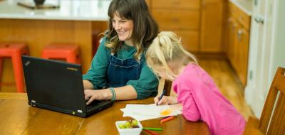 Mother on laptop happily doing accounting school work on laptop while young daughter colors next to her. 