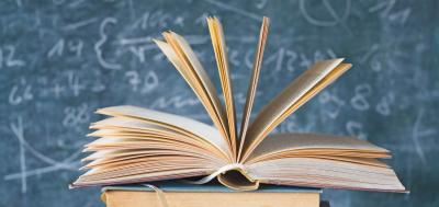 Stack of books, with top one open in front of a chalkboard