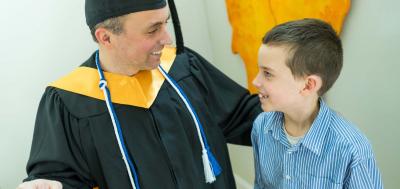 Male students in cap and gown smiling at young son