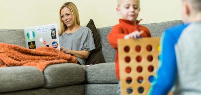 Adult female student doing schoolwork on couch while her children Connect Four game nearby