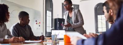 Woman of color standing, leading a meeting with a table full of employees