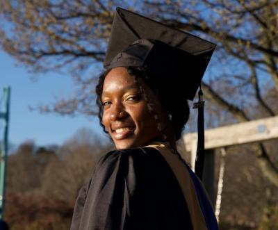 A female college graduate poses in front of large trees wearing a cap and gown.