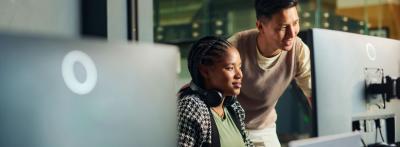 woman sitting and a man standing to her left both looking at two monitors