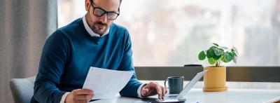 man with glasses and in blue sweater looking at a paper while typing on a laptop at his desk